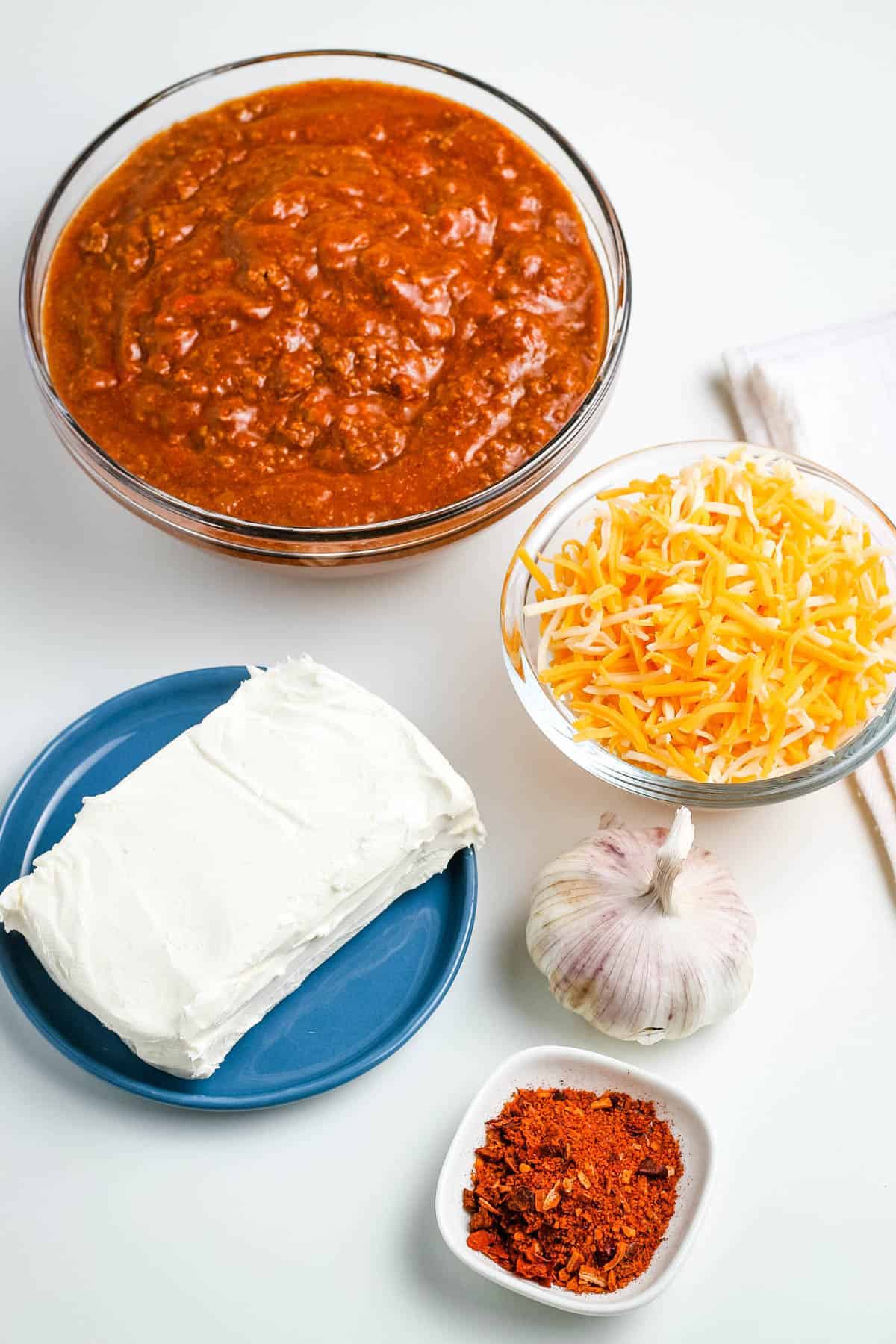 ingredients for chili dip in glass bowls, including chili without beans, shredded cheese, cream cheese, garlic, and taco seasoning