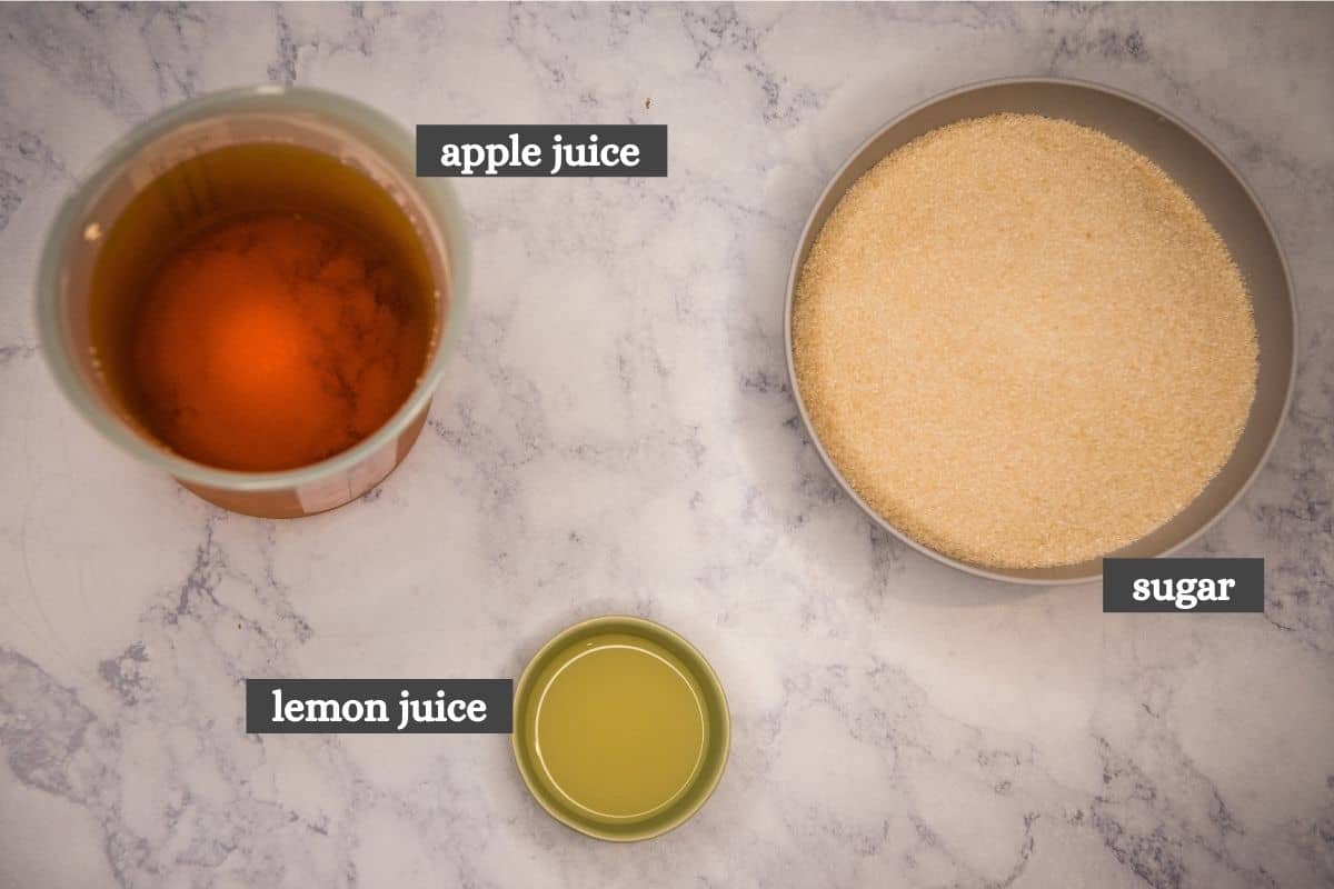 3 ingredients for apple jelly recipe without pectin, including apple juice, lemon juice, and sugar on white marble countertop
