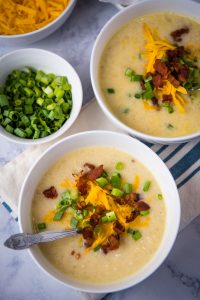 Instant Pot potato soup with bacon bits, cheddar cheese, and green onions in a small white bowl with a silver spoon on a white marbled countertop