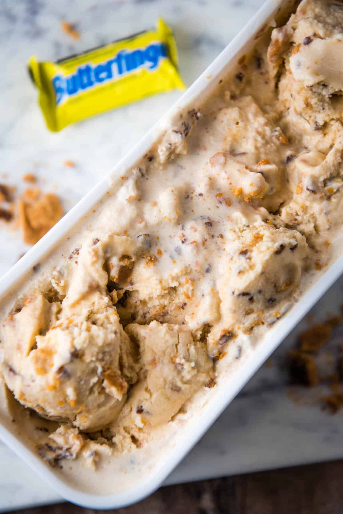 Butterfinger ice cream in white ice cream container next to candy bar on white marble countertop