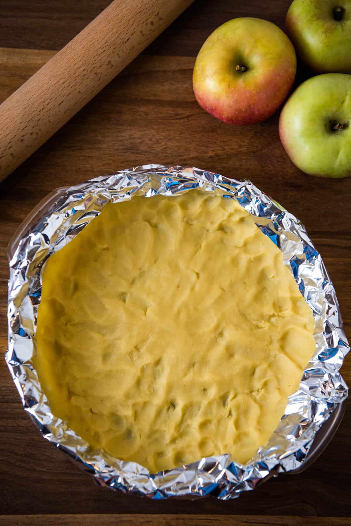 aluminum foil pie shield or foil protector over unbaked pie crust edges on wooden countertop with apples and rolling pin