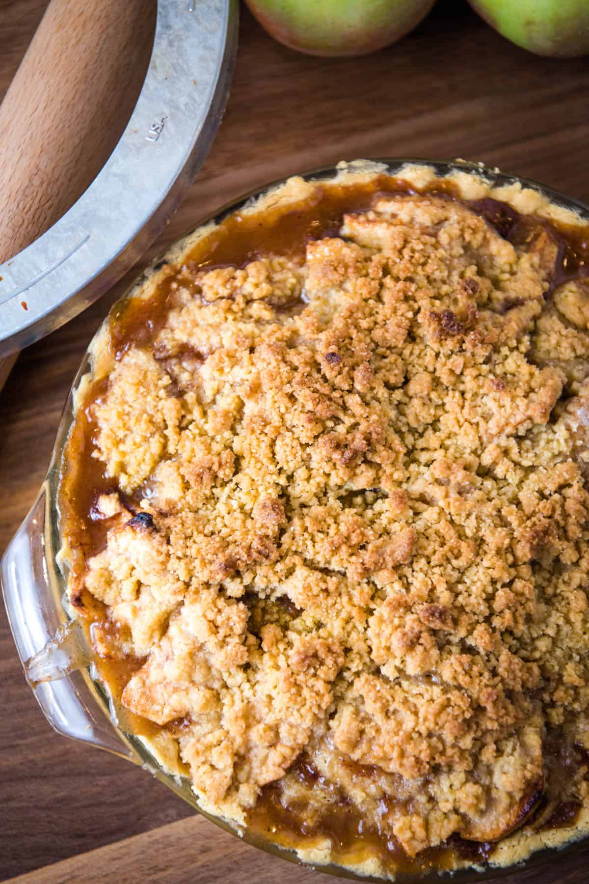 baked apple crumb pie with golden brown edges, pie shield, and rolling pin on wooden countertop