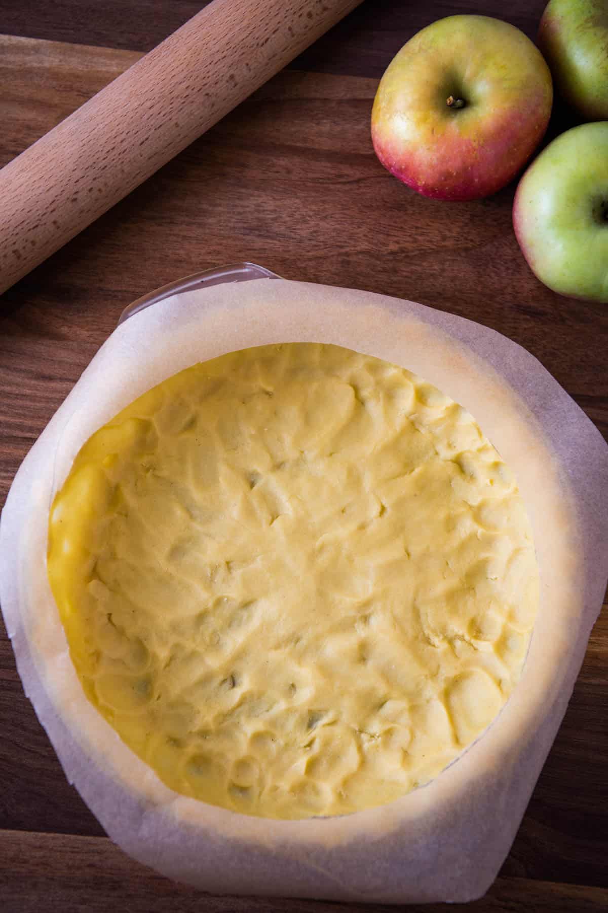 parchment paper pie shield on edges of unbaked pie crust on wooden countertop with apples and rolling pin