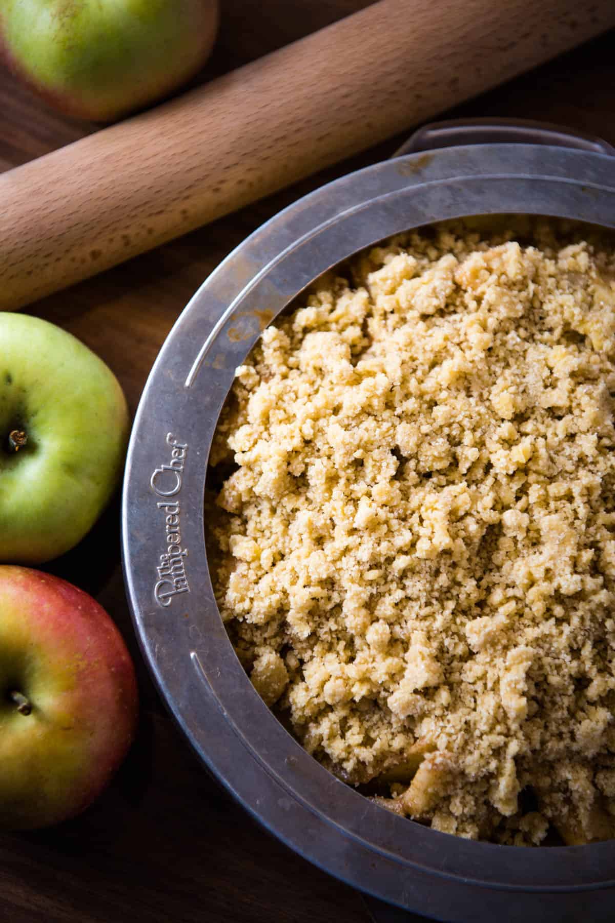 Pampered Chef pie crust protector over edges of apple crumb pie on wood countertop with rolling pin and apples