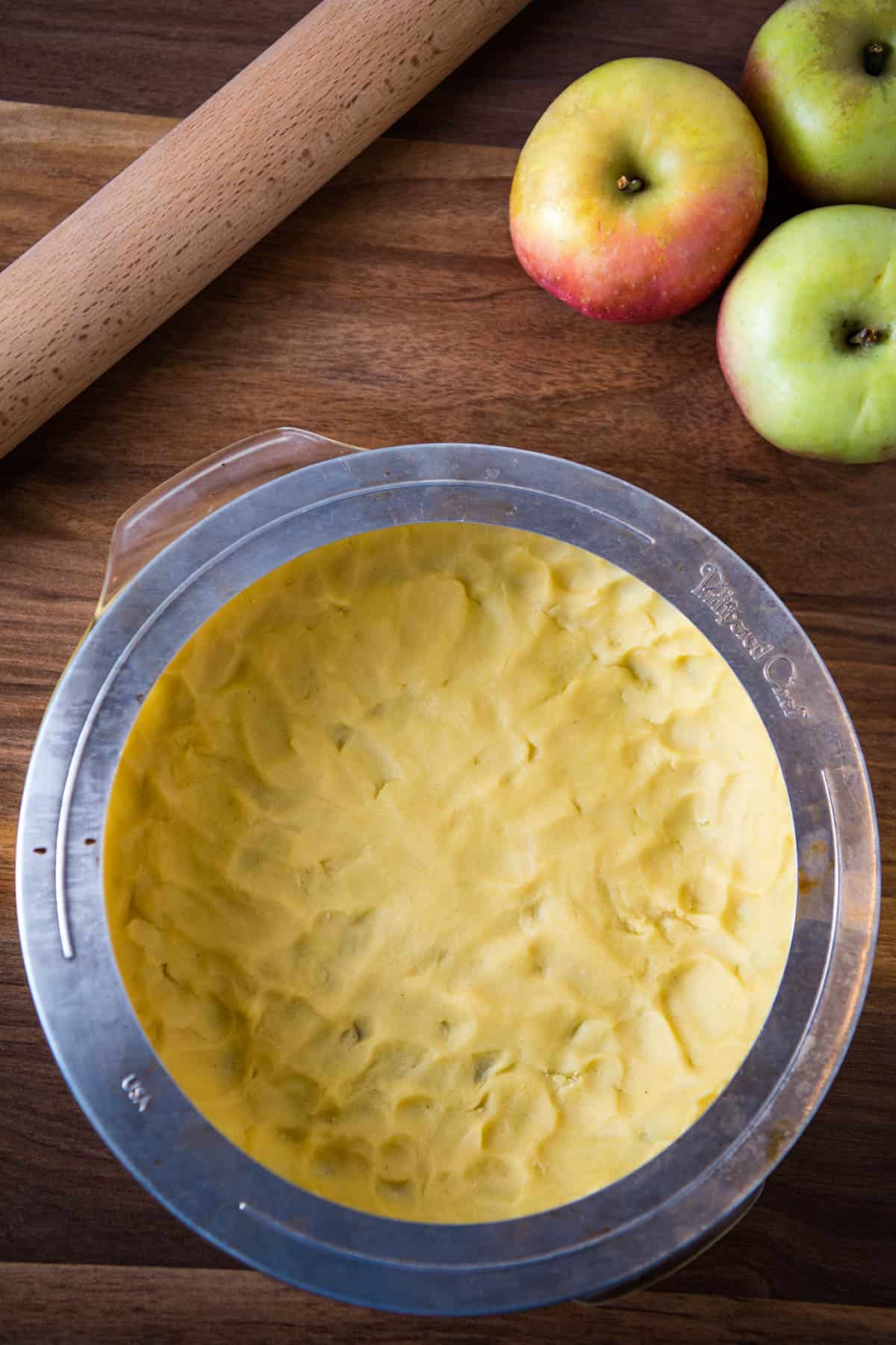 pie shield or pie crust protector on unbaked pie crust edges on wooden countertop with apples and wooden rolling pin