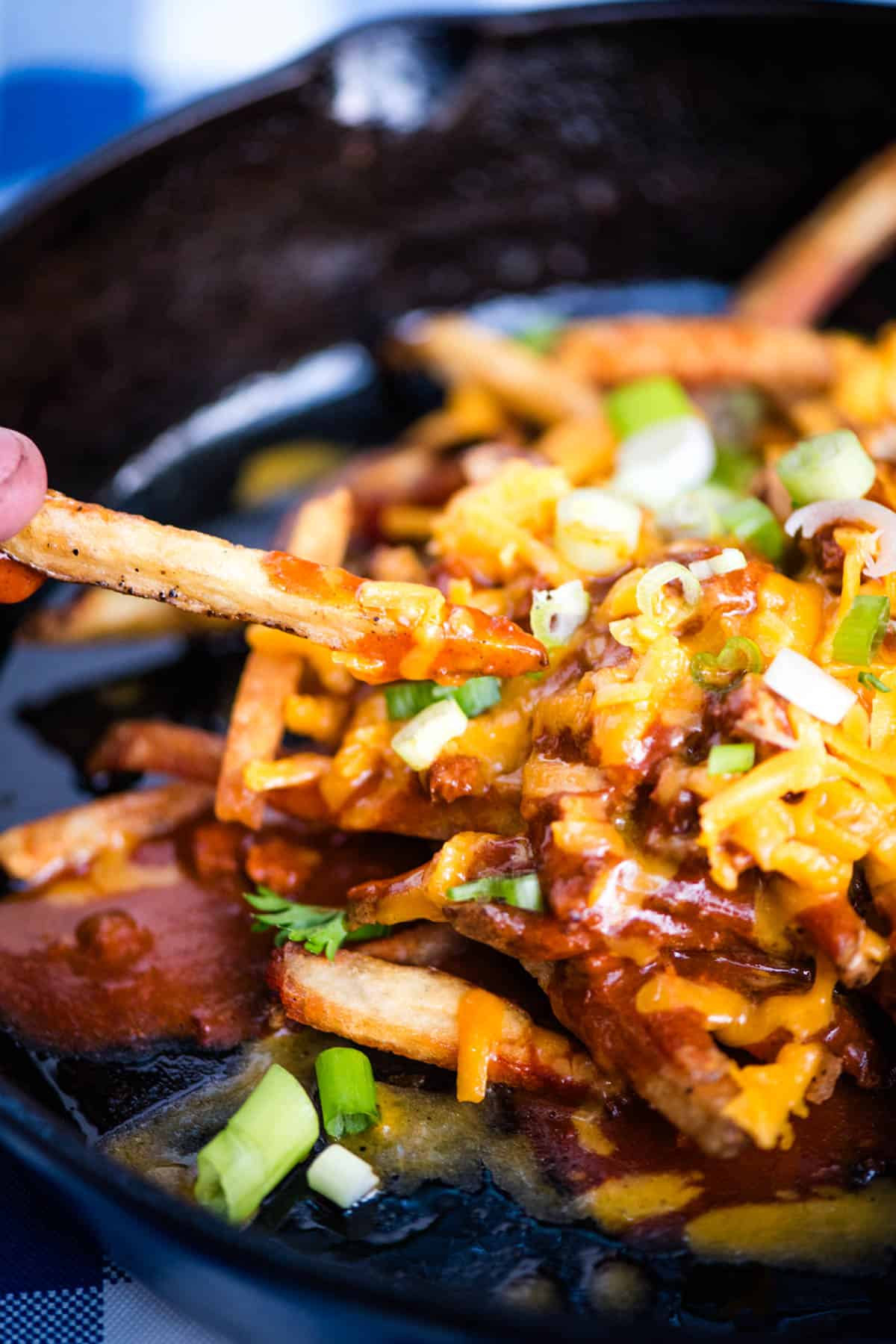 holding a crispy French fry over top of a large black cast iron skillet full of chili cheese fries