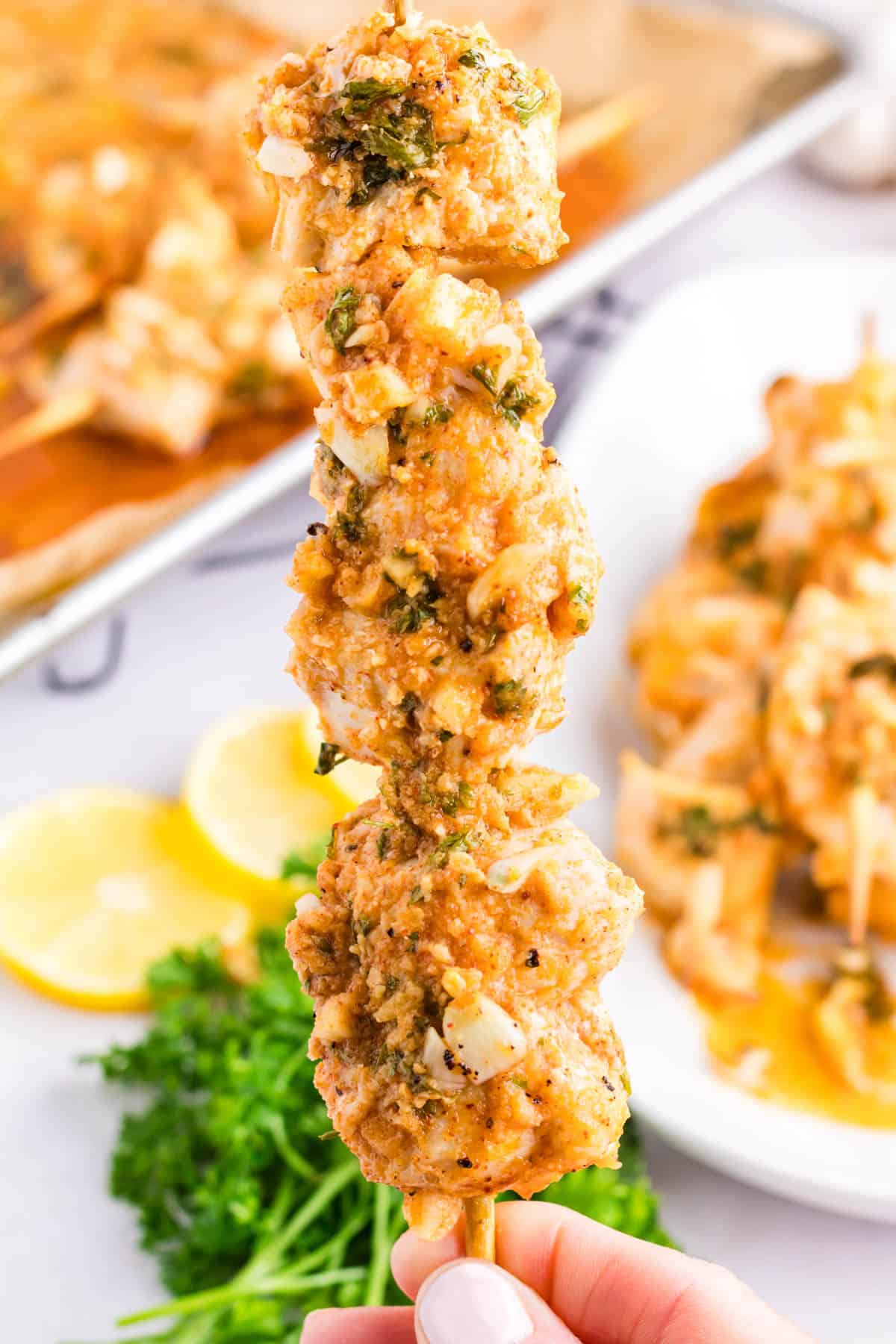 closeup photo of holding up a freshly baked garlic parmesan chicken kabob over a large silver baking sheet full of garlic parm chicken kabobs