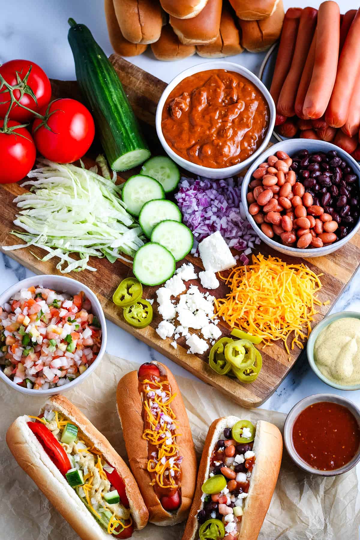 slow cooker hot dogs in hot dog buns next to all of the toppings lined out on a large wooden cutting board