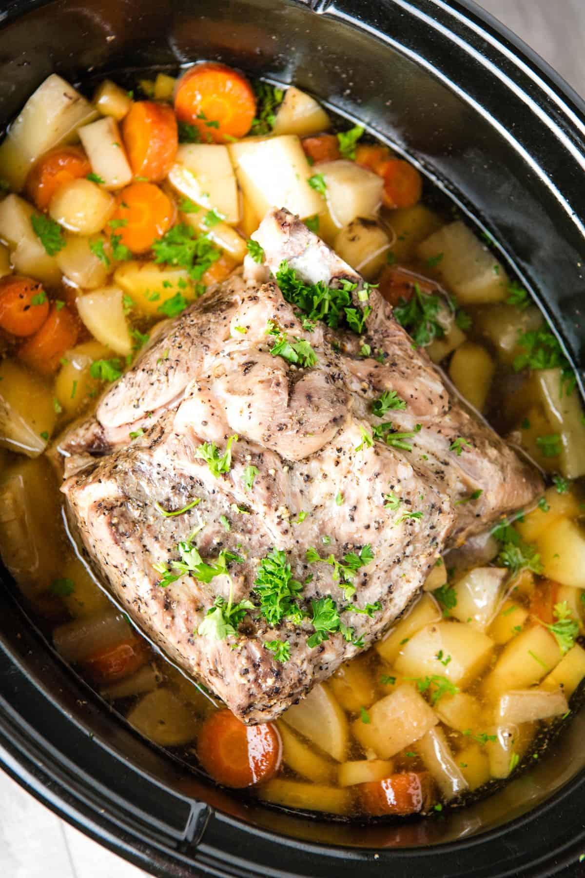 slow cooker pork roast on a bed of cooked root veggies with a garnish of fresh parsley in a large black slow cooker