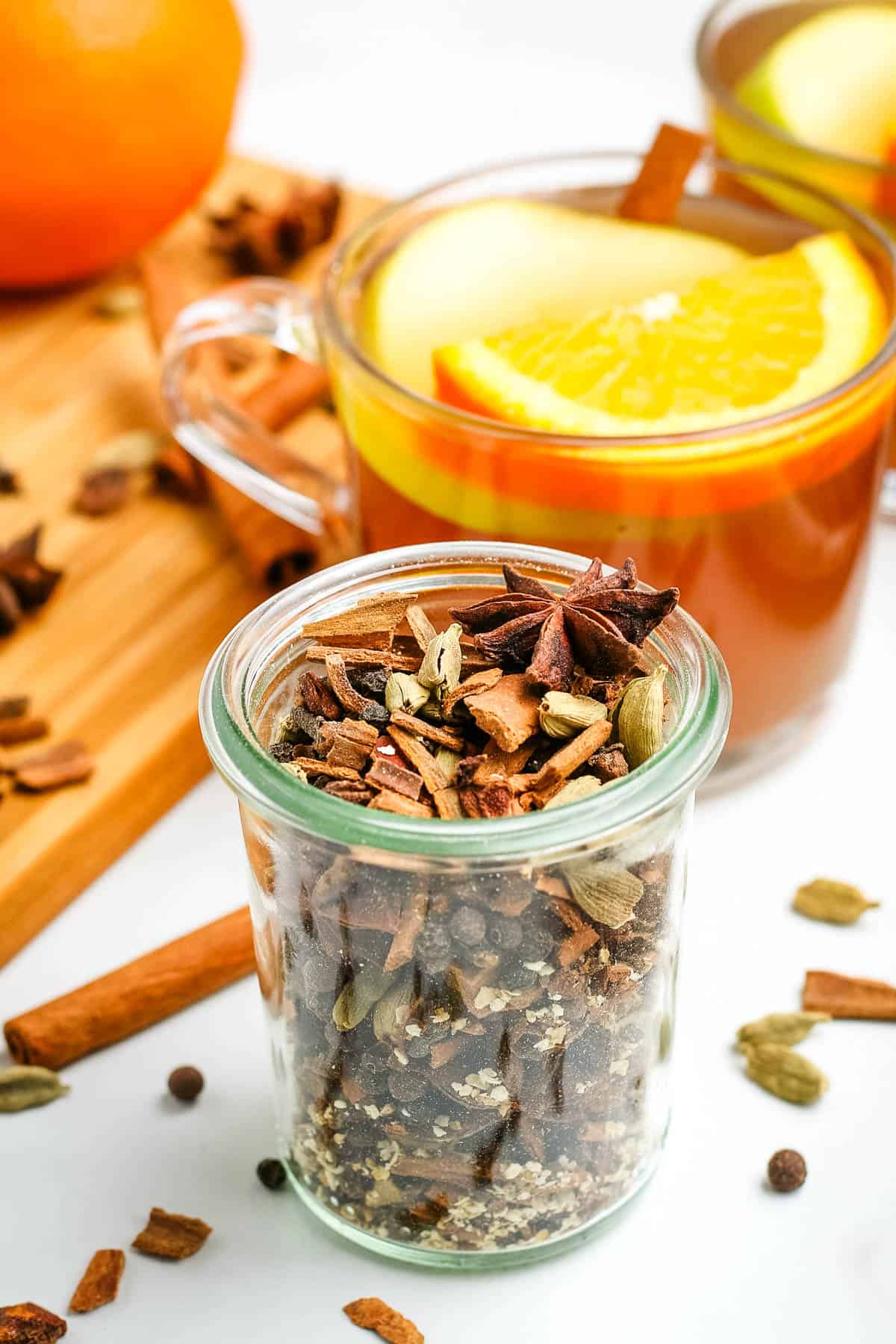 small glass jar full of mulling spices next to a small glass full of mulled cider with a slice of fresh lemon on top