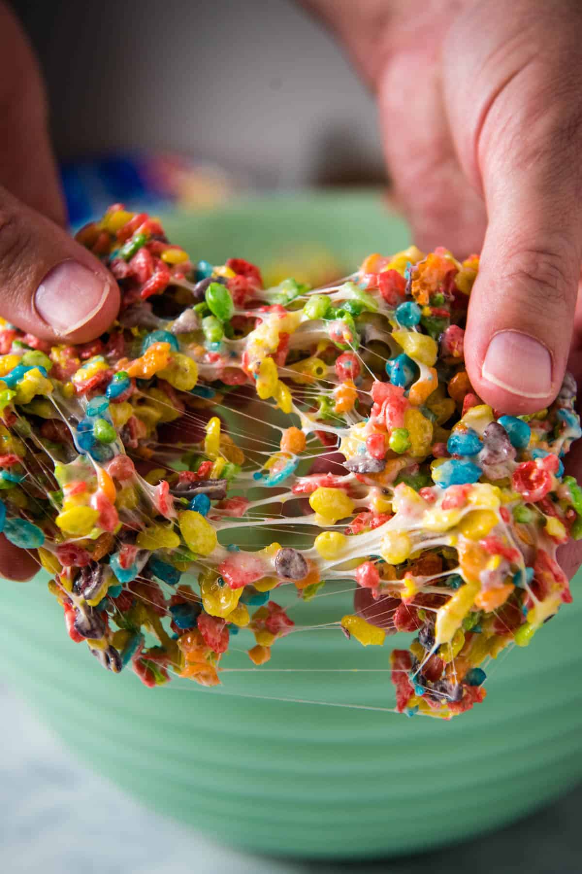 closeup shot showing pulling apart gooey fruity pebbles treats over top of a large green mixing bowl