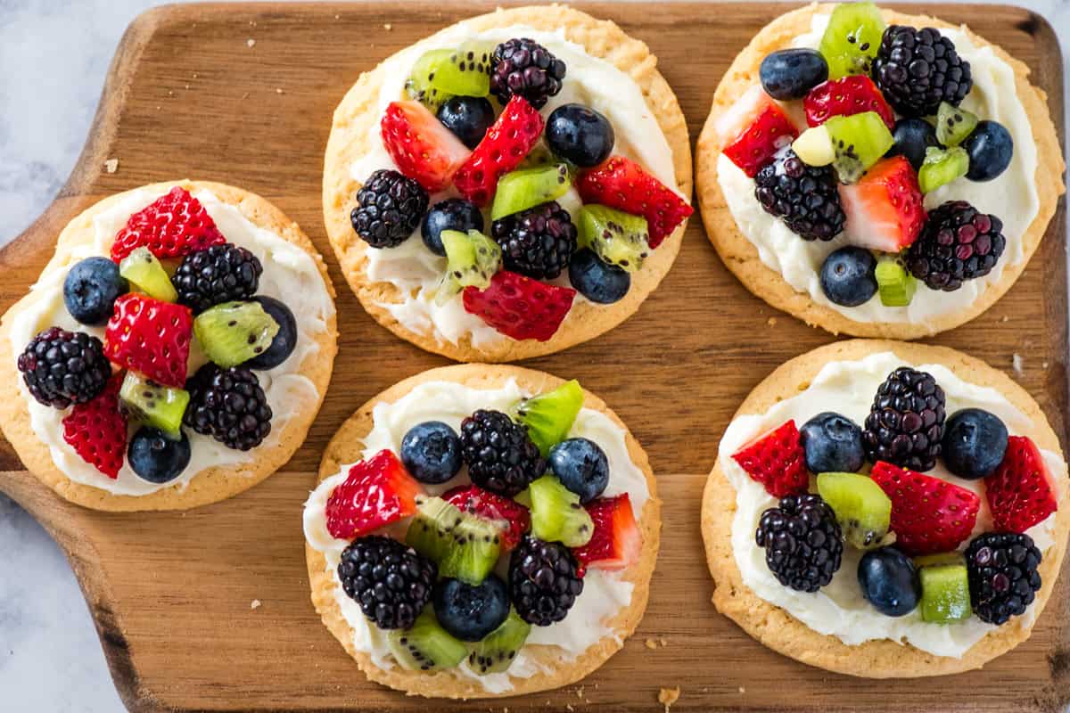 mini fruit pizza cookies lined out on a large brown wooden cutting board on a marbled white countertop
