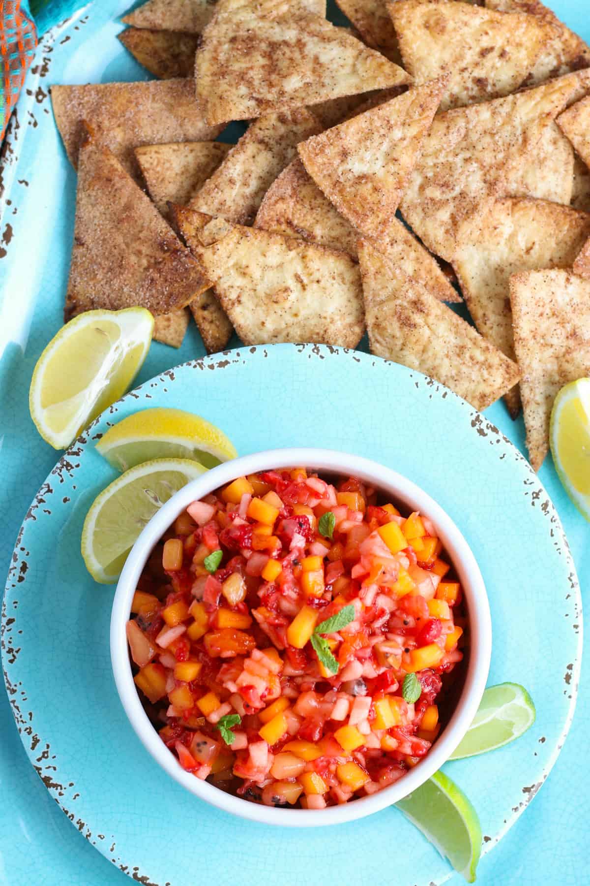 overhead photo showing fruit salsa in a white bowl on a decorative blue saucer with fresh lemon quarters and a pile of crispy cinnamon tortilla chips