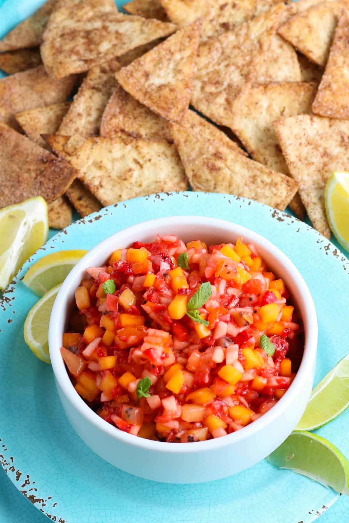 fruit salsa and cinnamon chips in a white bowl on a large blue decorative plate with fresh lemon quarters
